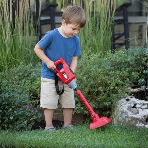 Dječak se igra s crvenom dječjom flaksericom - igračka trimer za travu u vrtu