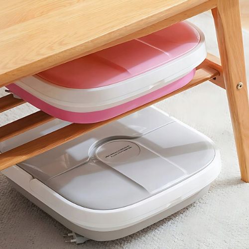 Ružičasti i sivi sklopivi masažeri za stopala potpuno sklopljeni na visinu od 10 cm i pospremljeni ispod drvene police radi uštede prostora.